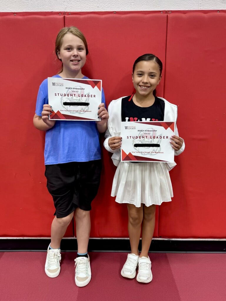 Two elementary school students standing together in front of a red wall, holding "REACH Ambassador - Student Leader" certificates with their names redacted.