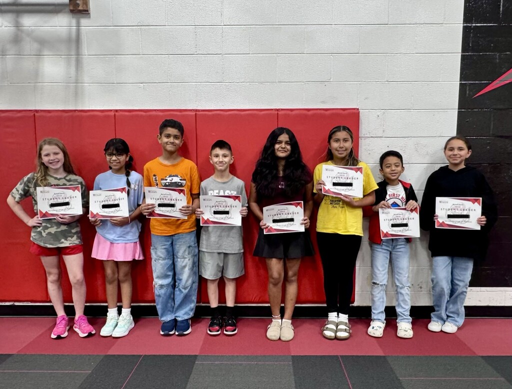 Eight elementary school students lined up in a gymnasium holding "Student Leader" certificates in front of a red wall.