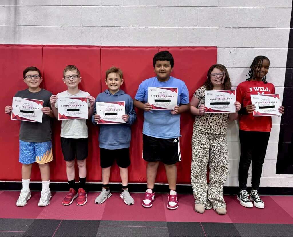 Six elementary school students standing in a line in front of a red wall in a gymnasium, each holding a certificate that reads "Student Leader."