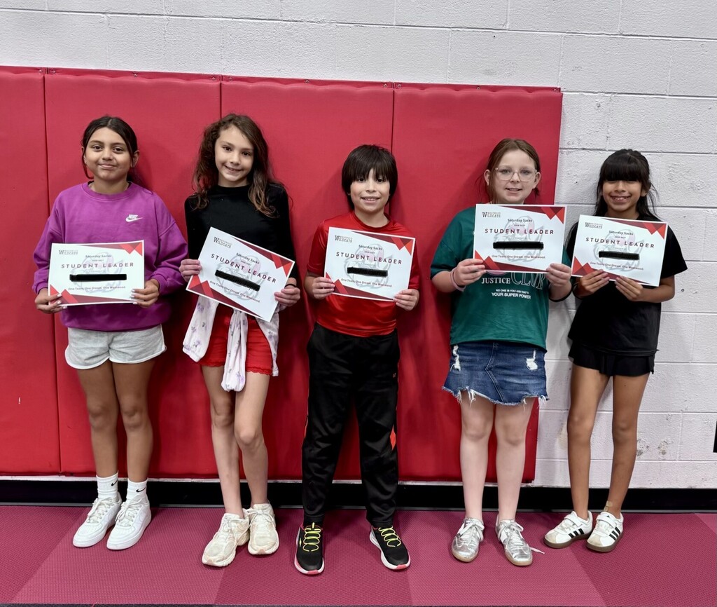 Five elementary school students standing together against a red padded wall, holding "Student Leader" certificates.