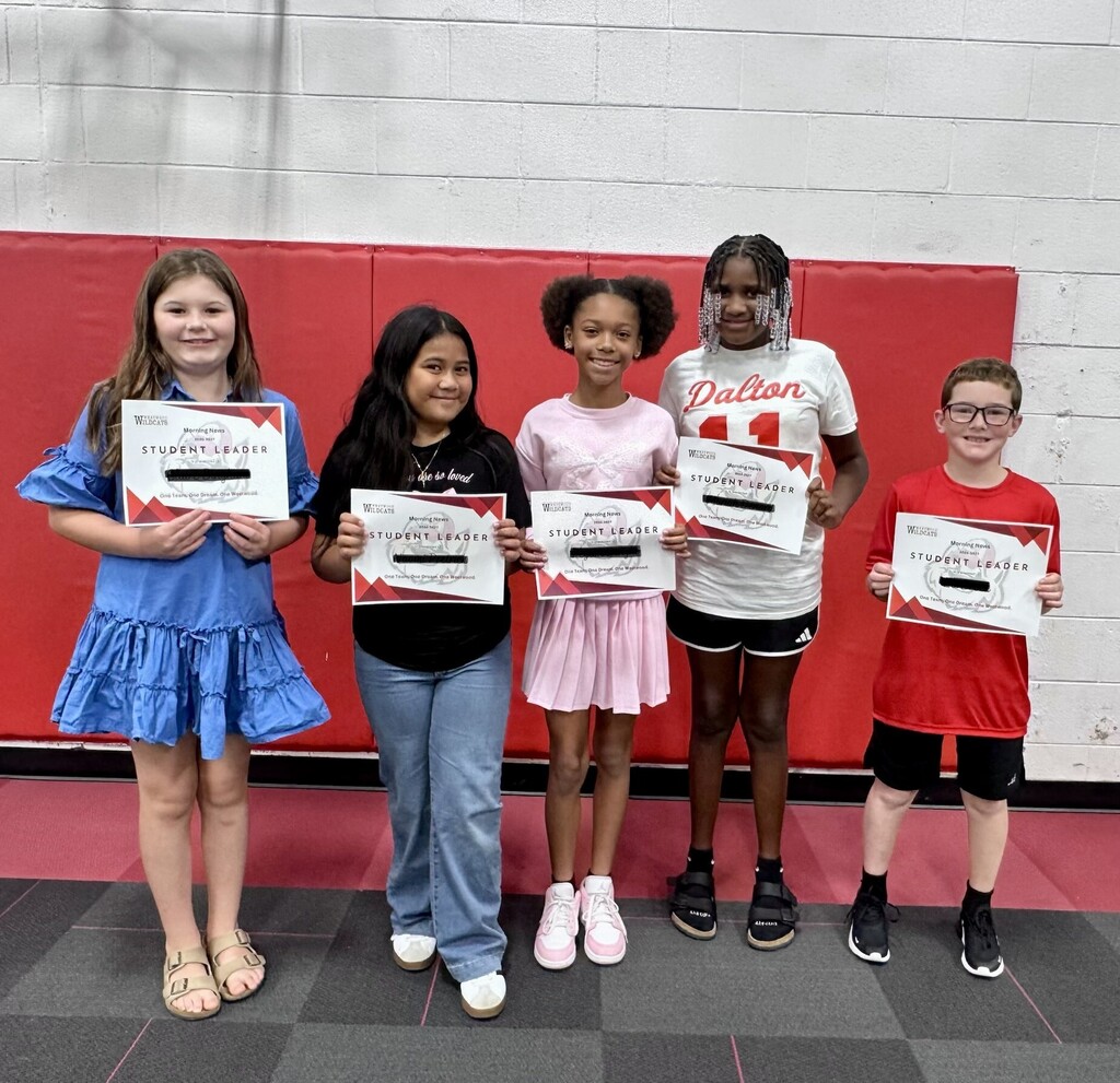 Five elementary school students standing side-by-side in a gymnasium in front of a red padded wall, each holding a "Student Leader" certificate with their name redacted.