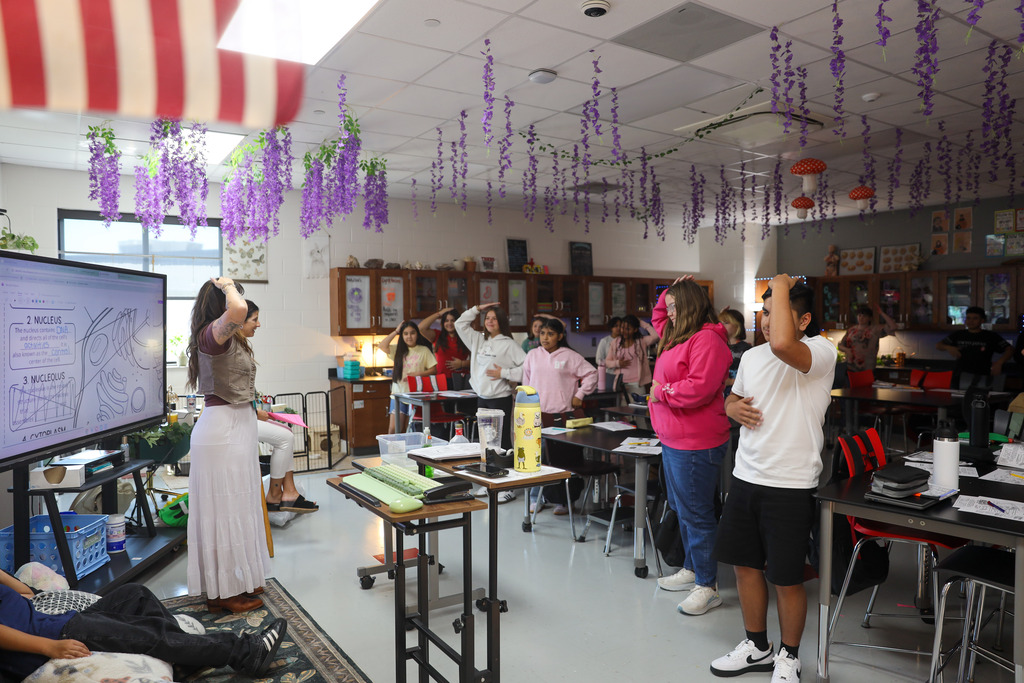 A female teacher stands at the front of a decorated classroom gesturing while speaking to her students. Seated at dark lab tables around her, middle school students look toward her attentively with notebooks and pencils out on their desks.
