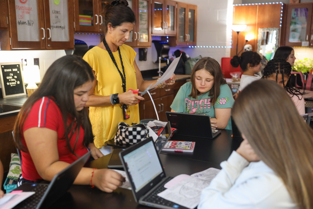 A female teacher wearing a yellow blouse and lanyard cuts paper with scissors while assisting a group of middle school students seated around a dark laboratory table. The students work on laptops and complete instructional worksheets, surrounded by science lab cabinetry.