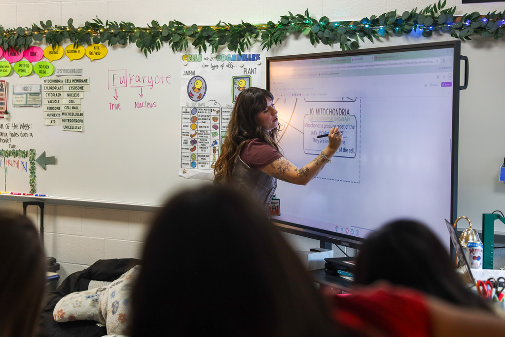 An interactive science classroom scene where a teacher in a white skirt and her students all stand up placing their hands on their heads to demonstrate an educational concept or movement. Hanging purple wisteria vines decorate the entire classroom ceiling.