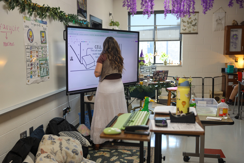 A science teacher with long dark hair and tattoos writes on a digital smartboard displaying a diagram about cell organelles, specifically "Mitochondria." A whiteboard to her left displays key cell terms such as "Eukaryote," "Nucleus," and "Cell Wall."