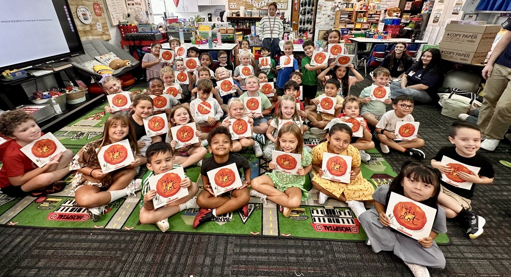 A large classroom filled with dozens of elementary students sitting cross-legged on a green play rug, each holding up a copy of the picture book The Dot by Peter H. Reynolds. Teachers and classroom aides sit and stand in the background near tables, storage shelves, and an interactive display screen.
