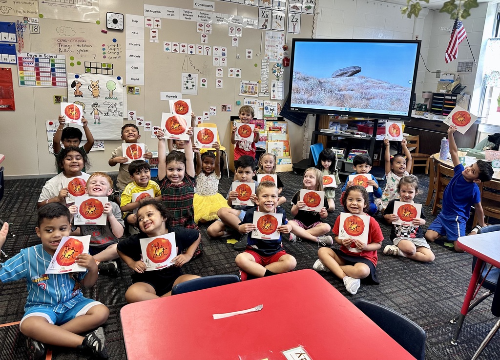 A large classroom filled with young students sitting cross-legged on the carpet, each holding up or displaying a copy of the picture book The Dot by Peter H. Reynolds. In the background, a large digital screen shows a landscape photograph of a rock formation under a bright sky. The surrounding walls are decorated with educational charts, including consonant and vowel references, calendar displays, and alphabet posters.