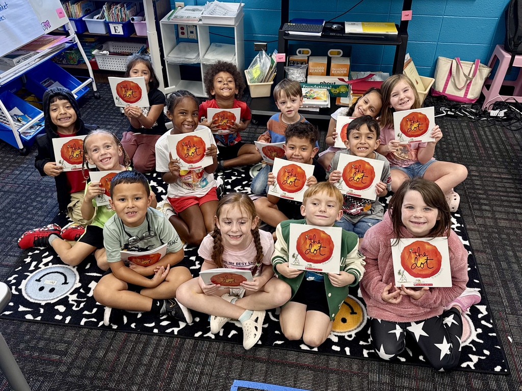 A group of 13 young students sitting closely together on a black-and-white pattern carpet in a classroom, each proudly displaying a copy of The Dot by Peter H. Reynolds. Four children sit in the front row, four sit directly behind them in the middle row, and five sit in the back row. Organized book bins, storage shelves, and classroom supplies line the blue wall in the background.