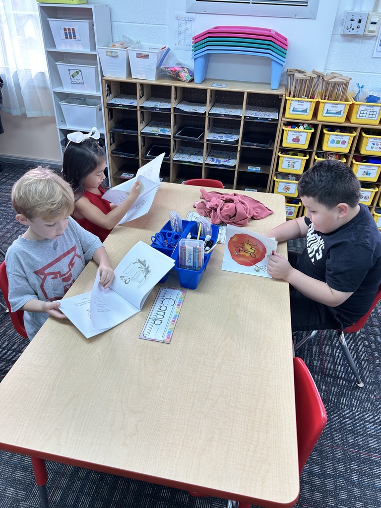 Three young children sitting around a light wooden classroom table, each reading or looking through a copy of The Dot by Peter H. Reynolds. A blue supply caddy filled with markers and scissors sits in the middle of the table alongside a printed nameplate reading "Camp." In the background, organized classroom storage shelves hold cubbies, bins, and colorful lap desks.
