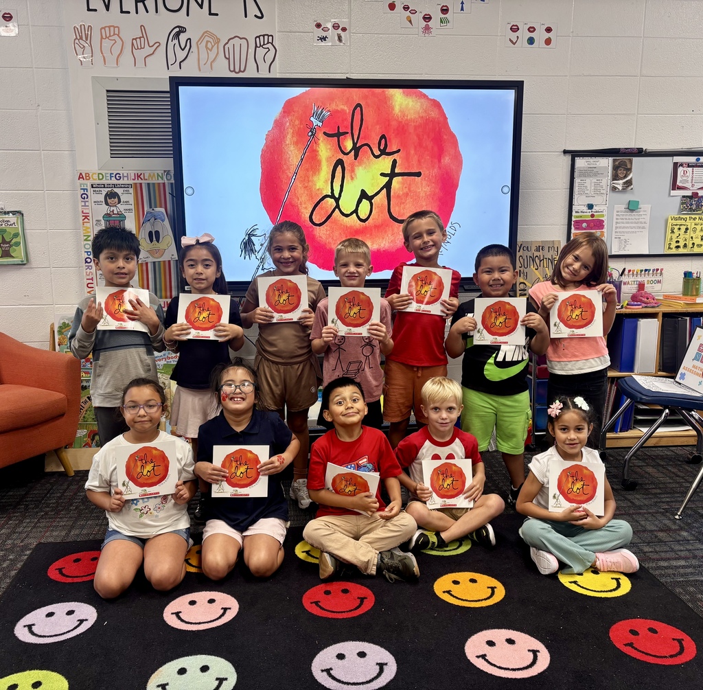 A group of 11 young students posing in a classroom while holding up copies of the picture book The Dot by Peter H. Reynolds. Five children sit cross-legged in the front row on a black rug decorated with colorful smiley faces, while six stand in a row behind them. In the center background, a large interactive monitor displays the main cover art of The Dot. The surrounding wall features educational posters, including sign language illustrations and alphabet charts.