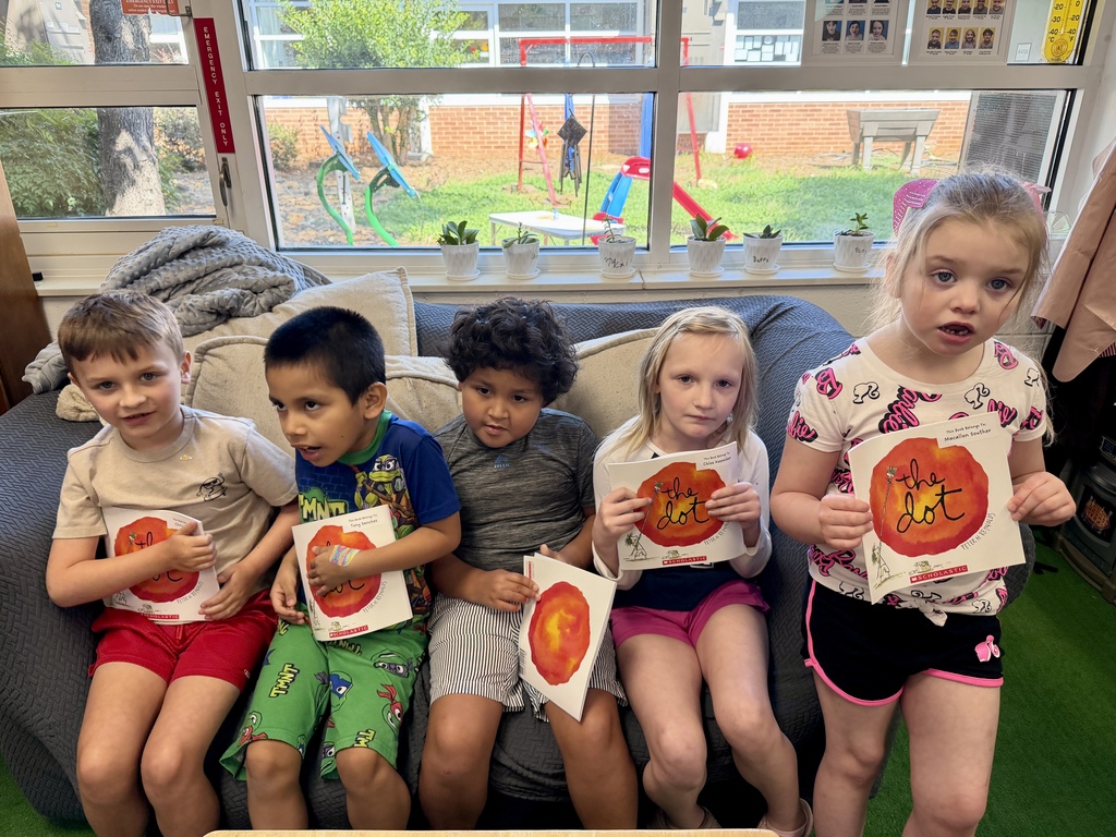 Five young children sitting together on a dark grey couch in a classroom, each holding a copy of The Dot by Peter H. Reynolds. Four of the children are seated side-by-side on the couch cushions, while a girl on the far right stands or leans close in a pink patterned t-shirt. Behind them, large windows look out onto an outdoor playground area, with small potted plants lined up along the windowsill.