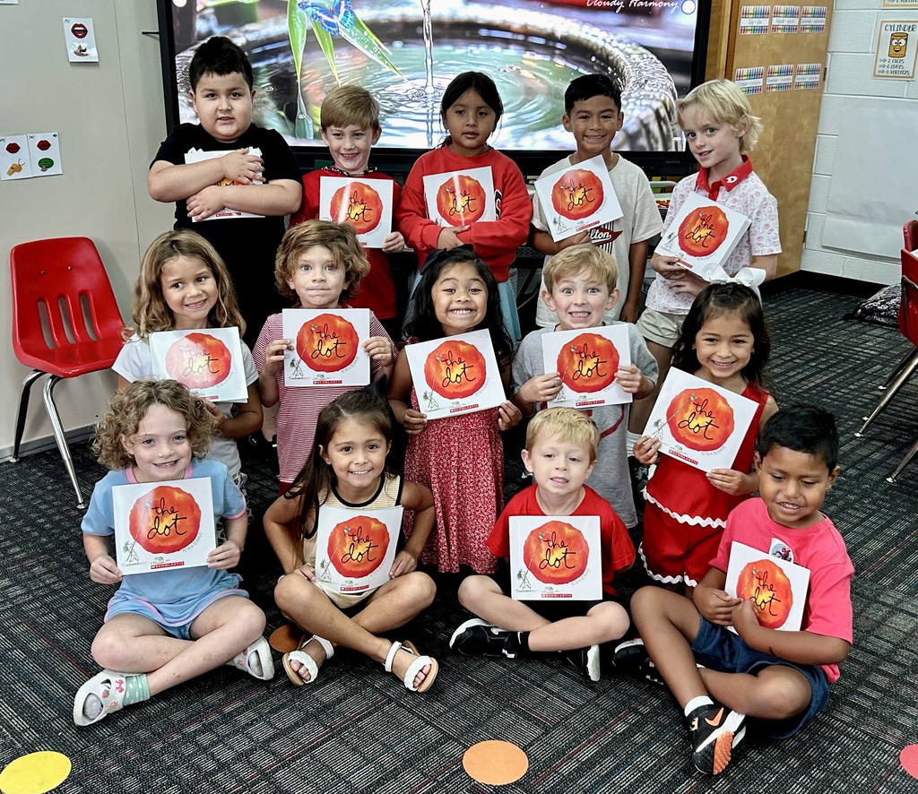 A group of 14 young students smiling while holding copies of the book The Dot by Peter H. Reynolds in a classroom. Four children sit cross-legged in the front row on a grey patterned carpet with colorful floor dots. Five children stand or sit in a middle row directly behind them, and five children stand in the back row. In the background, a large digital screen displays a serene water fountain image labeled "Cloudy Harmony."