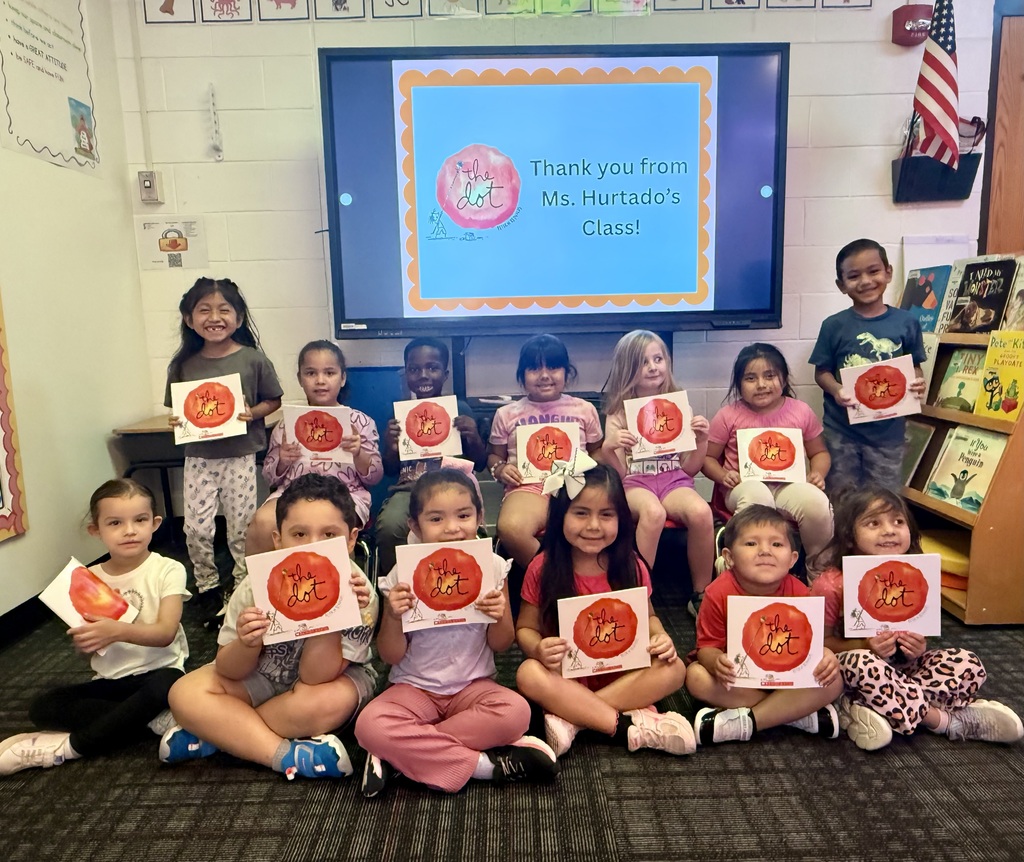 A group of 13 young children in a classroom smiling and holding up copies of the picture book The Dot by Peter H. Reynolds. Seven children are seated on the floor in the front row, while six are seated on chairs or standing behind them. In the center background, a large interactive display screen features a blue slide with an orange scalloped border reading "Thank you from Ms. Hurtado’s Class!" alongside The Dot book logo. To the right, a bookshelf displays various children's books beneath an American flag mounted on the wall.