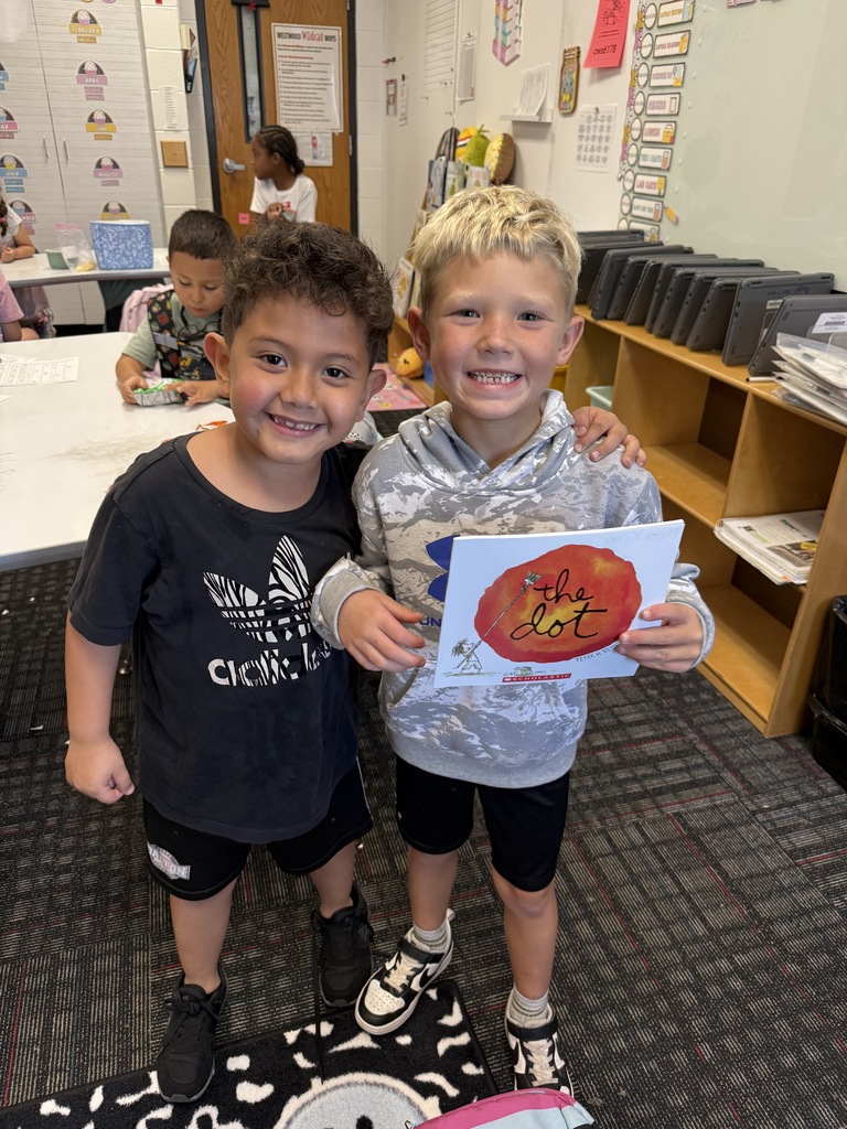 Two young boys smiling together in a classroom. The boy on the left has dark curly hair, wears a black Adidas t-shirt, and has his arm around his classmate. The boy on the right has short blonde hair, wears a grey patterned Under Armour hooded sweatshirt, and holds up a copy of the book The Dot by Peter H. Reynolds. In the background, other students sit at tables, and classroom shelves hold lined-up tablets and supplies