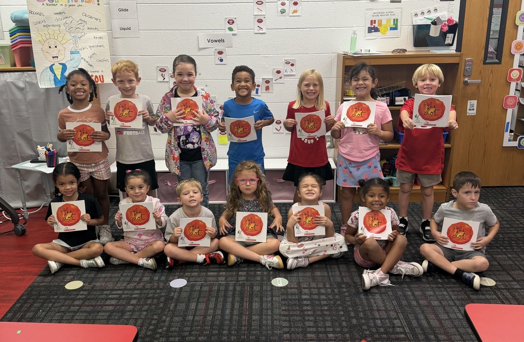A group of 14 young students smiling while holding up copies of the picture book The Dot by Peter H. Reynolds in a classroom. Seven children sit cross-legged in the front row on a dark patterned rug, while seven stand in a row behind them. In the background, educational posters including a "Scientist" chart and vowel references adorn the white brick wall, next to low wooden bookshelves and classroom cubbies.
