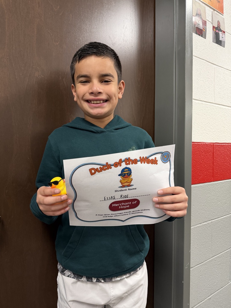 Student stands in a school hallway holding a Duck of the Week certificate and a small yellow duck award. The student is recognized by the Merchant of Hope team for encouraging others, spreading kindness, and helping classmates believe in themselves.