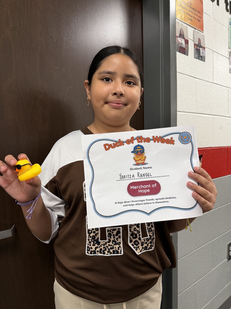 Student stands in a school hallway holding a Duck of the Week certificate and a small yellow duck award. The student is recognized by the Merchant of Hope team for encouraging others, spreading kindness, and helping classmates believe in themselves.