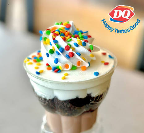 A Dairy Queen ice cream cupcake served in a clear plastic cup, featuring visible layers of chocolate and vanilla soft serve topped with a swirled peak of white icing and bright, multi-colored confetti sprinkles. The Dairy Queen logo with the slogan "Happy Tastes Good" appears in the top right corner.