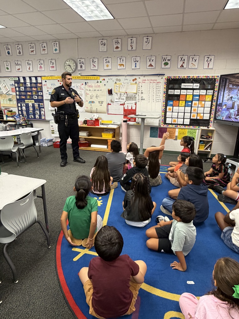 High-angle perspective showing a police officer standing in front of elementary students seated on a large blue classroom rug. The officer gestures with his hands while addressing the children, who face forward attentively near whiteboards and classroom displays.
