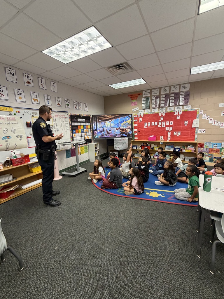 A police officer stands at the front of a primary classroom speaking to a group of young students gathered on a colorful area rug. The classroom is decorated with educational posters, an alphabet chart, and a digital display board.