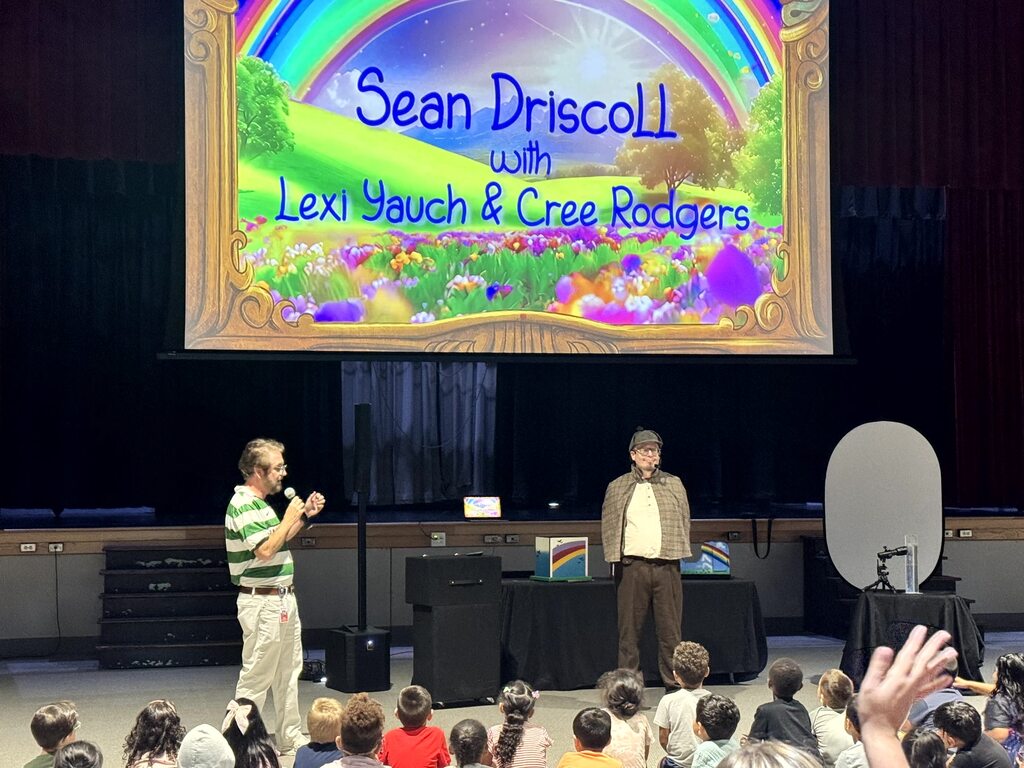 The images document an interactive performance for elementary school students featuring Sean Driscoll along with Lexi Yauch and Cree Rodgers.  Key Details from the Event  Performers: Sean Driscoll with Lexi Yauch & Cree Rodgers.  Attire: The main performer wears a Sherlock Holmes-inspired detective outfit, including a deerstalker cap, plaid cape, and round glasses.  Activities & Props:  Pouring liquid from a colorful, iridescent teapot into cups.  Handing or demonstrating a blue-and-yellow toy device with a young student helper on stage.  Performing puppetry with a blue and yellow spotted dragon/monster puppet.  Audience Interaction: The children sit on the auditorium floor, actively raising their hands and engaging with the presenter during the show.