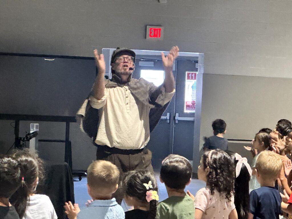 The images document an interactive performance for elementary school students featuring Sean Driscoll along with Lexi Yauch and Cree Rodgers.  Key Details from the Event  Performers: Sean Driscoll with Lexi Yauch & Cree Rodgers.  Attire: The main performer wears a Sherlock Holmes-inspired detective outfit, including a deerstalker cap, plaid cape, and round glasses.  Activities & Props:  Pouring liquid from a colorful, iridescent teapot into cups.  Handing or demonstrating a blue-and-yellow toy device with a young student helper on stage.  Performing puppetry with a blue and yellow spotted dragon/monster puppet.  Audience Interaction: The children sit on the auditorium floor, actively raising their hands and engaging with the presenter during the show.