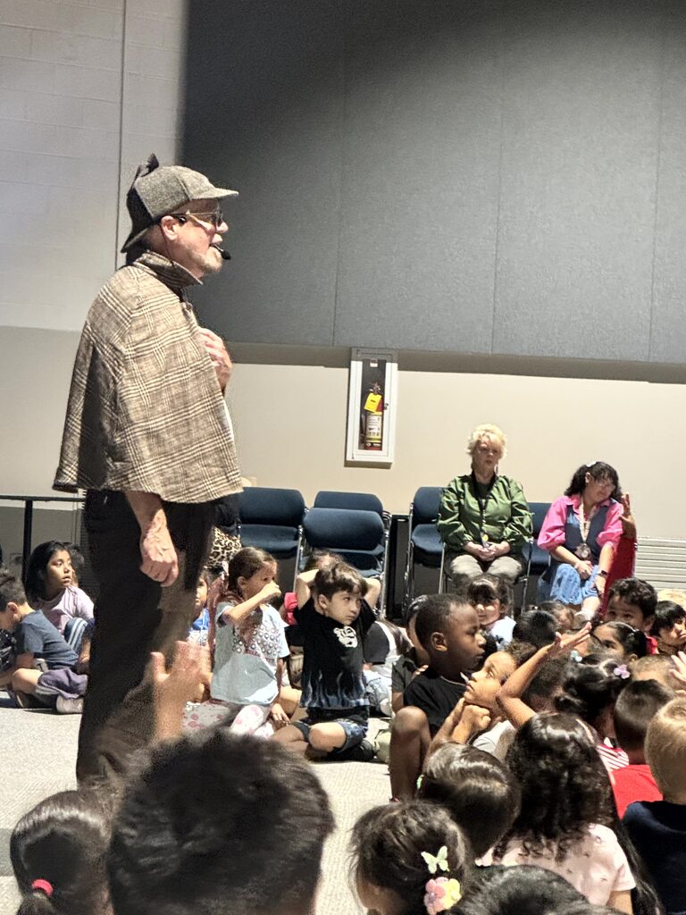 The images document an interactive performance for elementary school students featuring Sean Driscoll along with Lexi Yauch and Cree Rodgers.  Key Details from the Event  Performers: Sean Driscoll with Lexi Yauch & Cree Rodgers.  Attire: The main performer wears a Sherlock Holmes-inspired detective outfit, including a deerstalker cap, plaid cape, and round glasses.  Activities & Props:  Pouring liquid from a colorful, iridescent teapot into cups.  Handing or demonstrating a blue-and-yellow toy device with a young student helper on stage.  Performing puppetry with a blue and yellow spotted dragon/monster puppet.  Audience Interaction: The children sit on the auditorium floor, actively raising their hands and engaging with the presenter during the show.