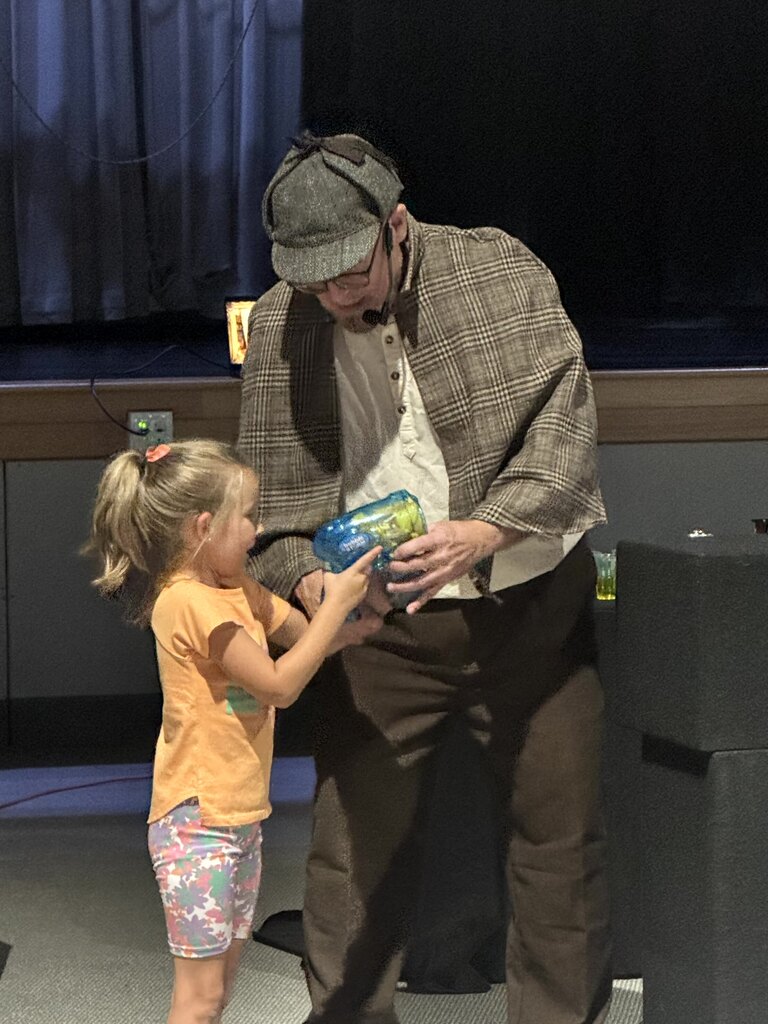 The images document an interactive performance for elementary school students featuring Sean Driscoll along with Lexi Yauch and Cree Rodgers.  Key Details from the Event  Performers: Sean Driscoll with Lexi Yauch & Cree Rodgers.  Attire: The main performer wears a Sherlock Holmes-inspired detective outfit, including a deerstalker cap, plaid cape, and round glasses.  Activities & Props:  Pouring liquid from a colorful, iridescent teapot into cups.  Handing or demonstrating a blue-and-yellow toy device with a young student helper on stage.  Performing puppetry with a blue and yellow spotted dragon/monster puppet.  Audience Interaction: The children sit on the auditorium floor, actively raising their hands and engaging with the presenter during the show.