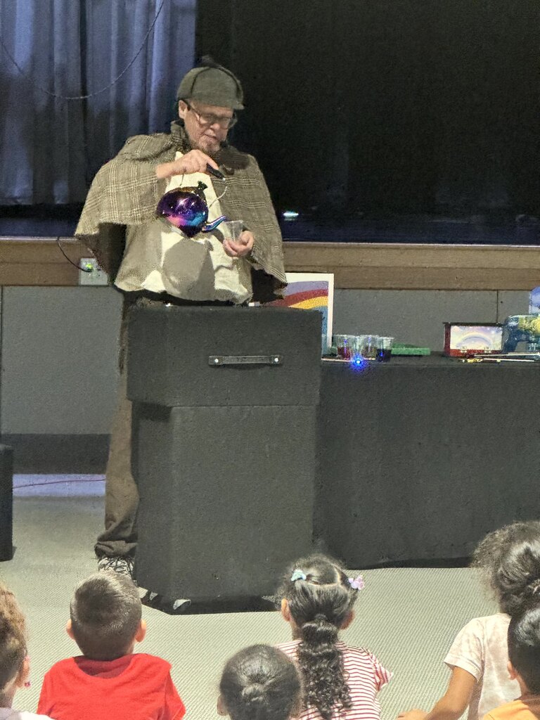 The images document an interactive performance for elementary school students featuring Sean Driscoll along with Lexi Yauch and Cree Rodgers.  Key Details from the Event  Performers: Sean Driscoll with Lexi Yauch & Cree Rodgers.  Attire: The main performer wears a Sherlock Holmes-inspired detective outfit, including a deerstalker cap, plaid cape, and round glasses.  Activities & Props:  Pouring liquid from a colorful, iridescent teapot into cups.  Handing or demonstrating a blue-and-yellow toy device with a young student helper on stage.  Performing puppetry with a blue and yellow spotted dragon/monster puppet.  Audience Interaction: The children sit on the auditorium floor, actively raising their hands and engaging with the presenter during the show.