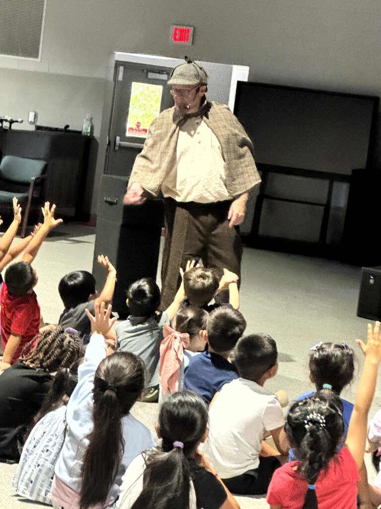 The images document an interactive performance for elementary school students featuring Sean Driscoll along with Lexi Yauch and Cree Rodgers.  Key Details from the Event  Performers: Sean Driscoll with Lexi Yauch & Cree Rodgers.  Attire: The main performer wears a Sherlock Holmes-inspired detective outfit, including a deerstalker cap, plaid cape, and round glasses.  Activities & Props:  Pouring liquid from a colorful, iridescent teapot into cups.  Handing or demonstrating a blue-and-yellow toy device with a young student helper on stage.  Performing puppetry with a blue and yellow spotted dragon/monster puppet.  Audience Interaction: The children sit on the auditorium floor, actively raising their hands and engaging with the presenter during the show.