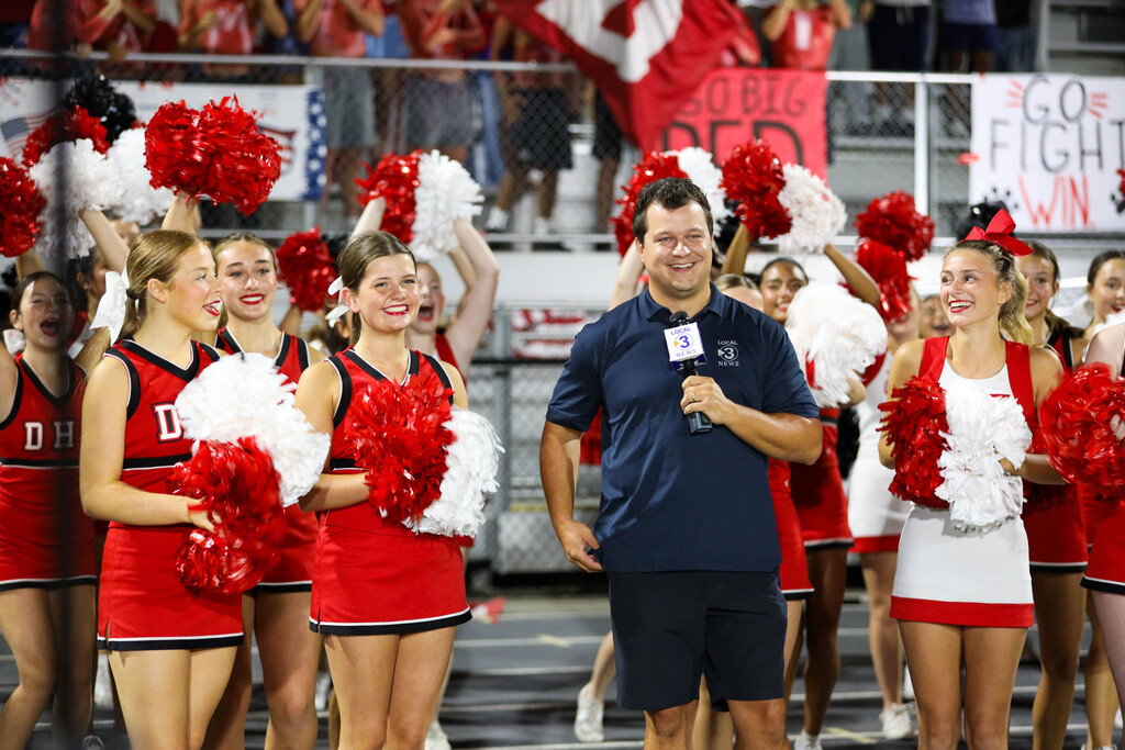A Local 3 News reporter holding a microphone while standing in front of cheerleaders.