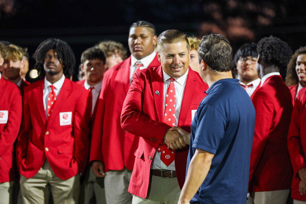 A news reporter shaking hands with a Dalton coach in a red blazer surrounded by players.