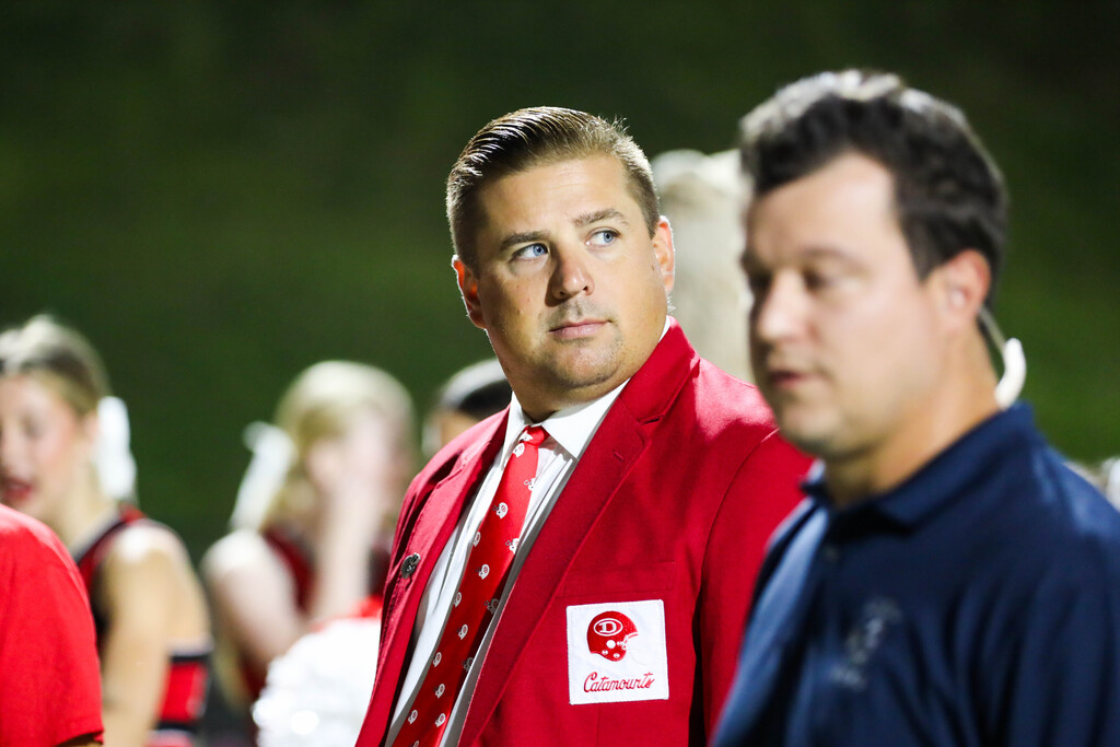 A coach in a red blazer with a Catamounts patch looking across the field next to a news reporter