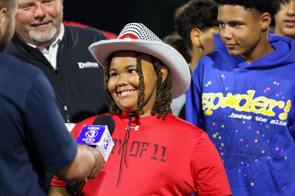 A young student wearing a cowboy hat smiling during an on-field TV news interview.