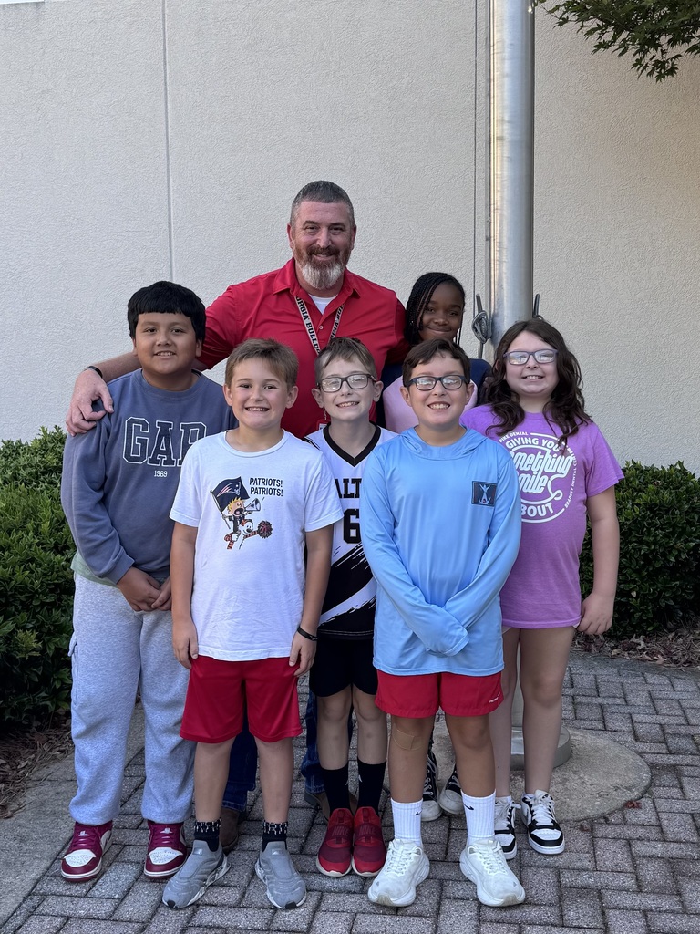 An adult man in a red shirt stands with six smiling children outside next to a flagpole.