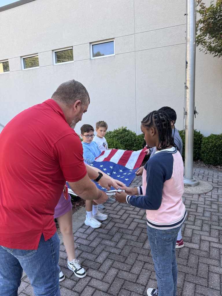 An adult man in a red polo shirt guides four children outside as they hold and carefully fold an American flag near a flagpole.