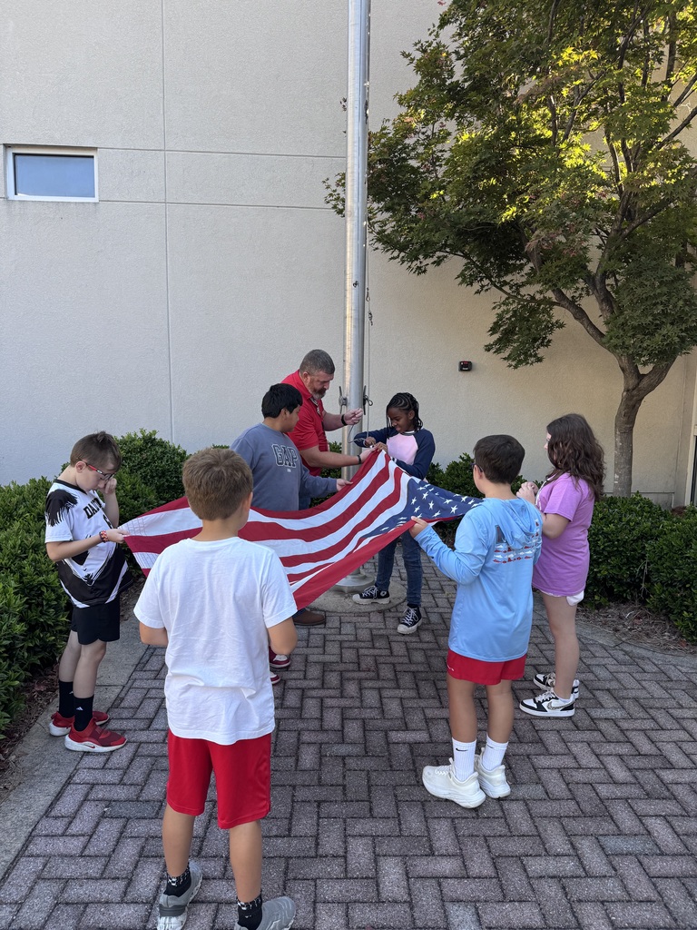 An adult man in a red shirt guides a group of children as they hold an American flag stretched open near a tall silver flagpole outside.