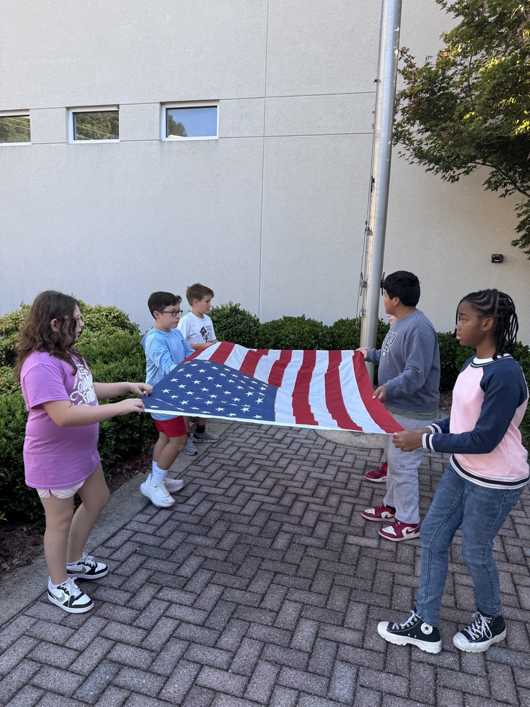 A group of five children hold an American flag stretched flat outdoors near a flagpole.