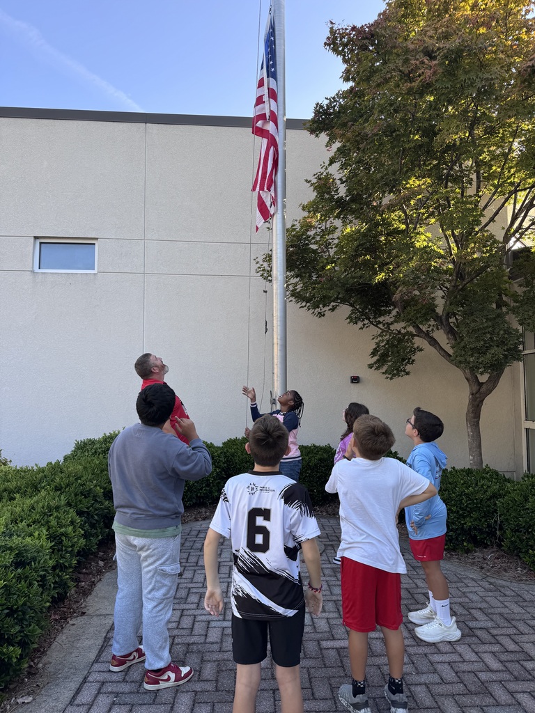 An adult and a group of children look up as an American flag is raised on a metal flagpole outside.
