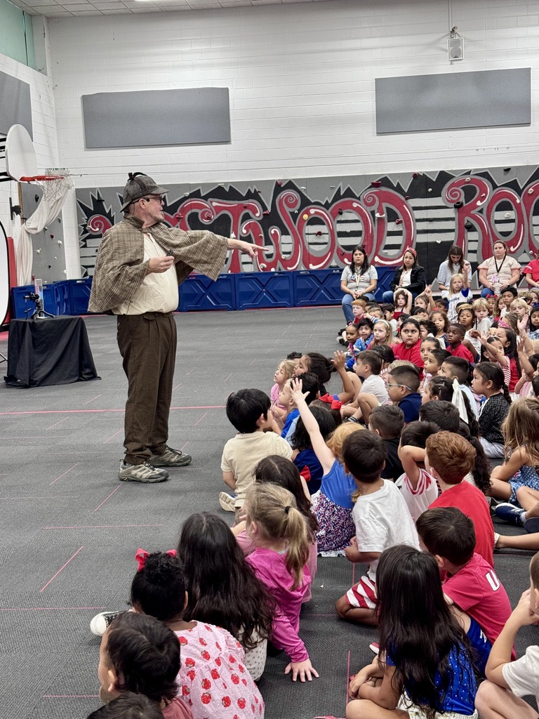 A performer in a detective costume points outward while presenting to a large audience of children sitting on a gymnasium floor.