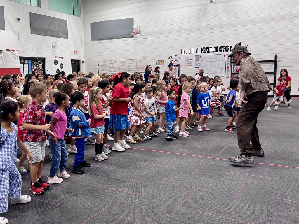 A performer in a detective costume moves animatedly in front of a large line of elementary students gathered on a school gymnasium floor.