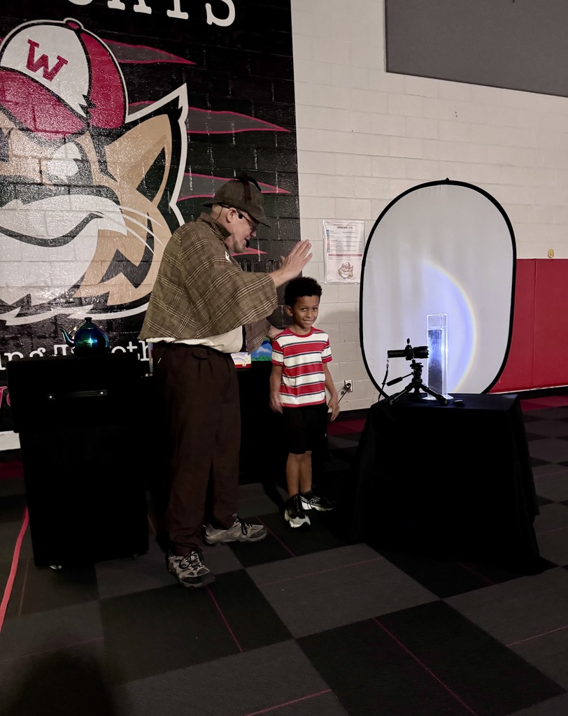 A performer dressed as a detective gestures toward a smiling young boy standing on stage next to a light demonstration setup.
