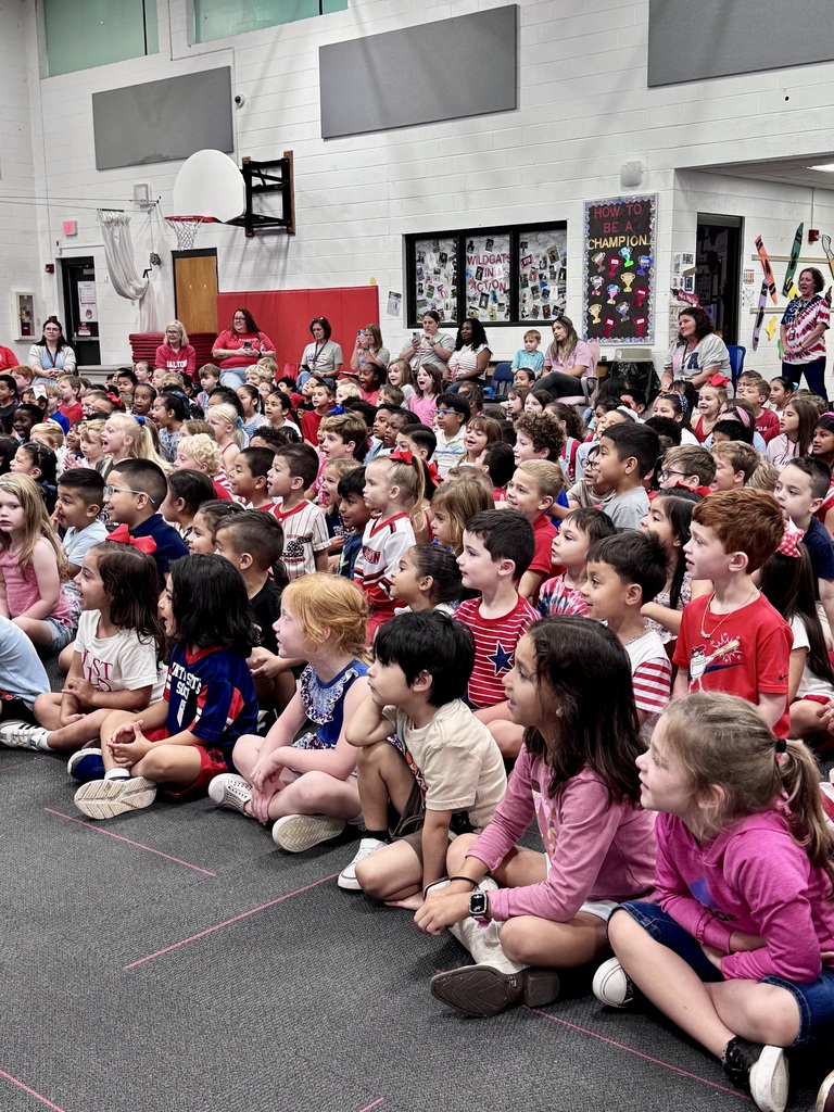 A large group of elementary school students sit cross-legged on a gymnasium floor watching a presentation.