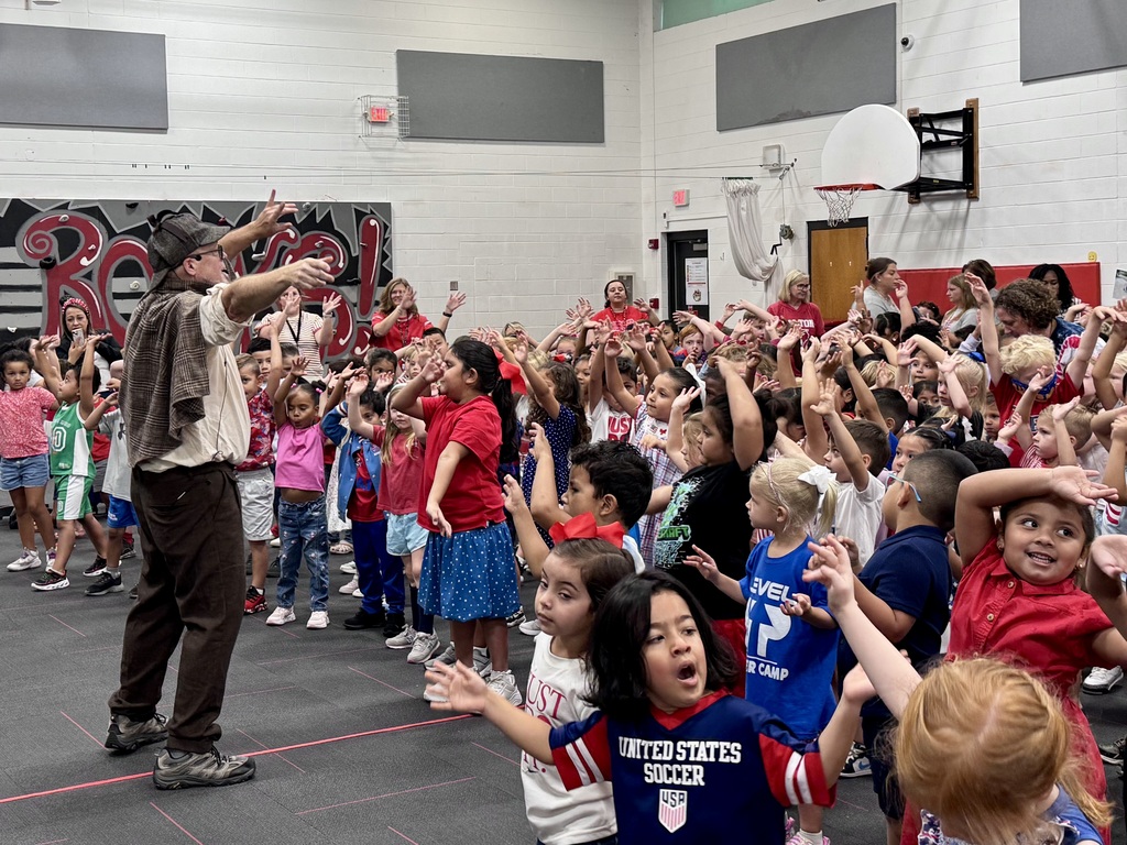 A performer in a detective costume leads a gymnasium full of elementary students with raised hands during an interactive assembly.