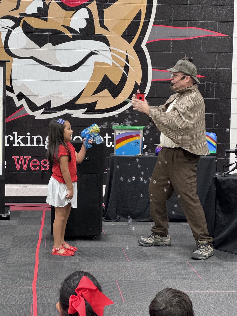A performer dressed as a detective interacts with a young girl who is shooting a bubble gun during a school assembly.