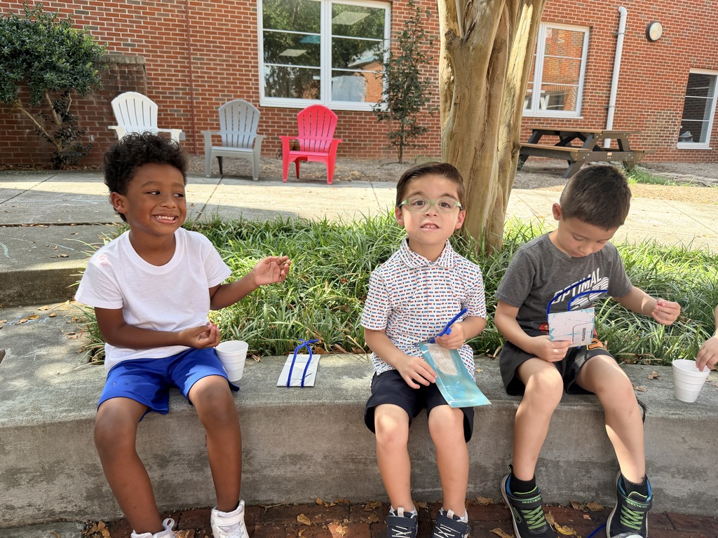 Young students are working on a hands-on outdoor craft activity in a courtyard area.  **Key Details**  * **Activity**: The children are assembling custom craft cards using blue pipe cleaners strung with red and white beads. * **Setting**: An outdoor courtyard adjacent to a brick school building, featuring concrete benches, trees, and colorful patio chairs. * **Highlights**: * Children working on the pavement and sitting along a concrete retaining wall. * Students posing together to proudly display their completed bead-and-pipe-cleaner crafts.