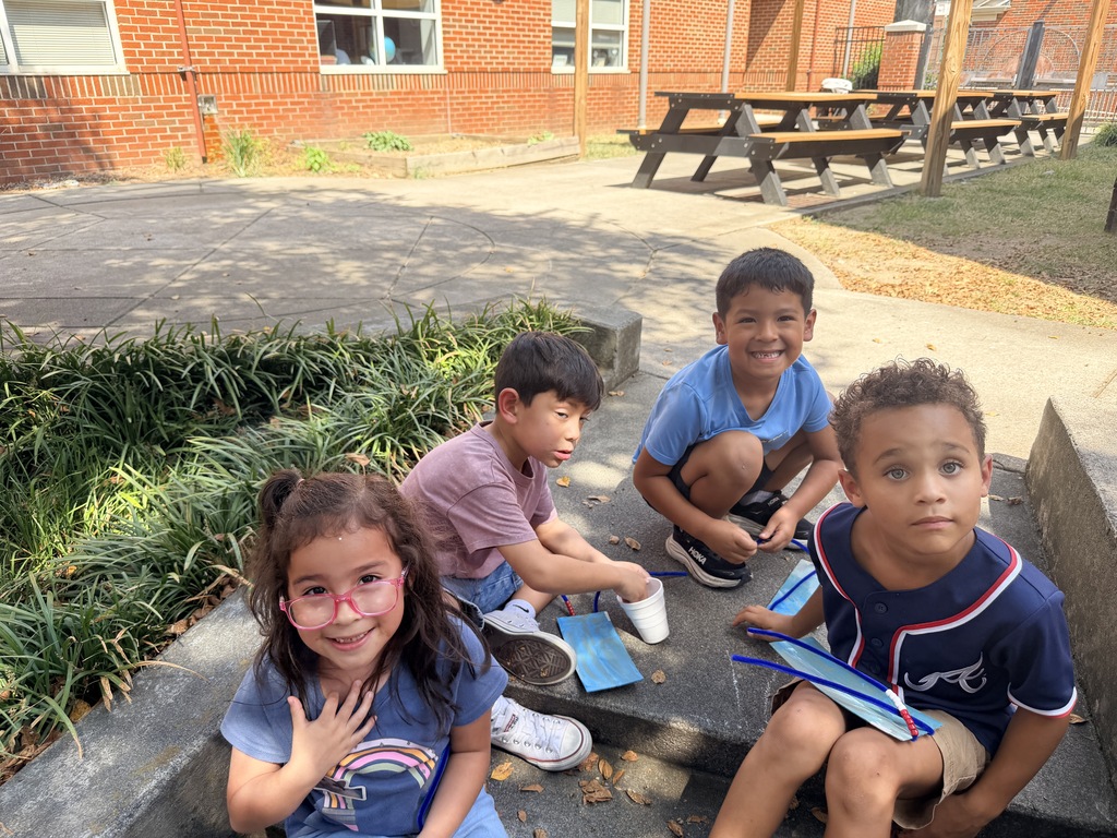 Four students sitting together on courtyard steps, smiling and holding their craft materials while enjoying an outdoor activity.