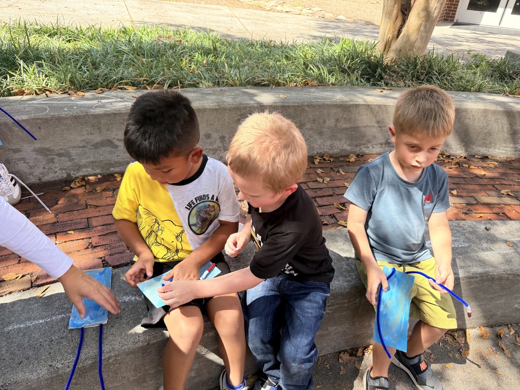 Three young boys sitting on a concrete step outdoors, leaning toward each other as they work together on their blue pipe cleaner and card crafts.