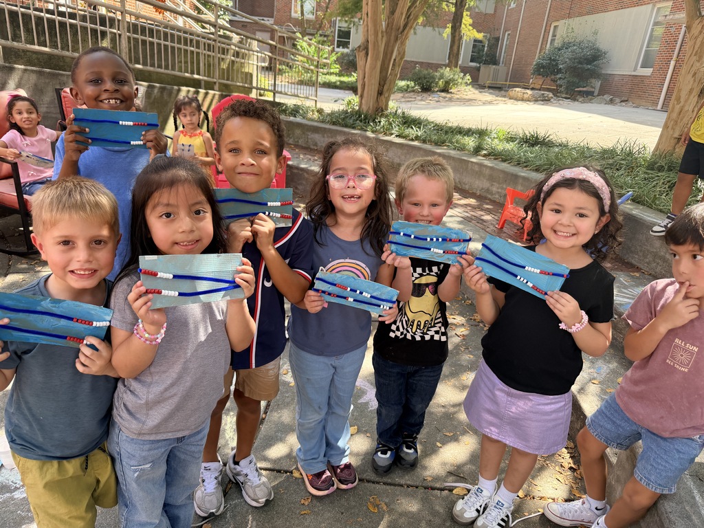 Young students are working on a hands-on outdoor craft activity in a courtyard area.  **Key Details**  * **Activity**: The children are assembling custom craft cards using blue pipe cleaners strung with red and white beads. * **Setting**: An outdoor courtyard adjacent to a brick school building, featuring concrete benches, trees, and colorful patio chairs. * **Highlights**: * Children working on the pavement and sitting along a concrete retaining wall. * Students posing together to proudly display their completed bead-and-pipe-cleaner crafts.