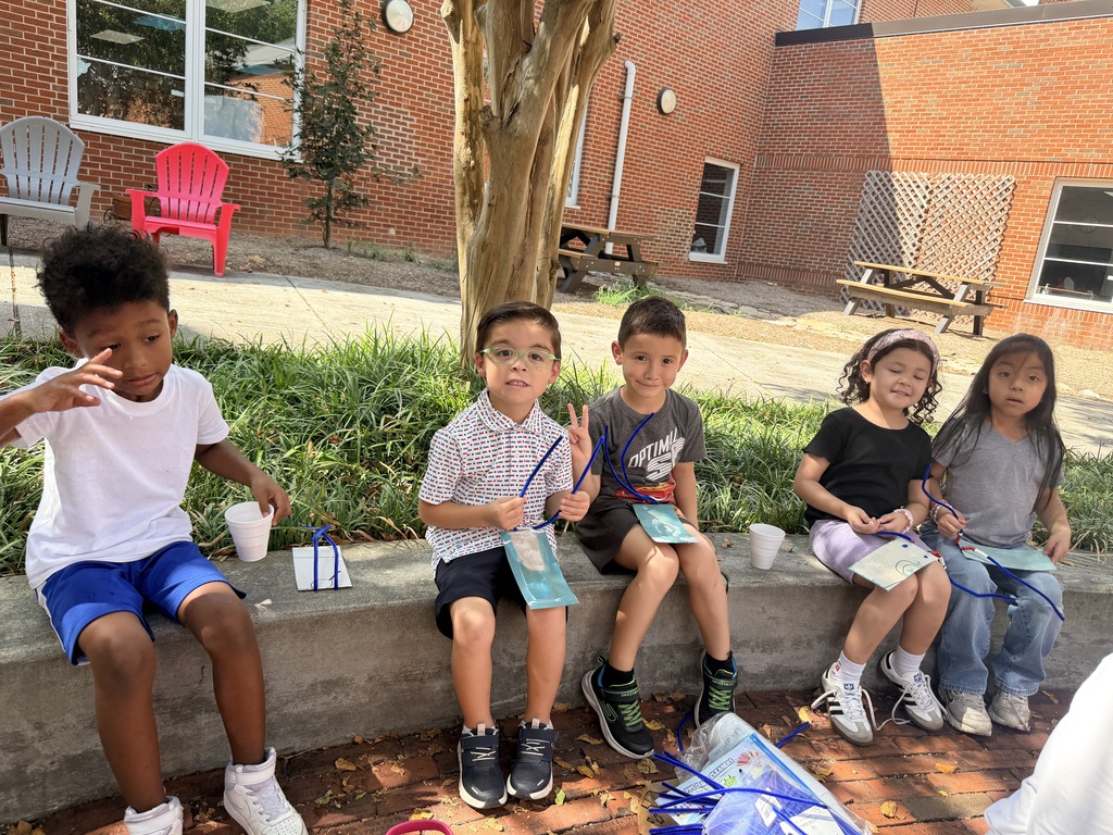Young students are working on a hands-on outdoor craft activity in a courtyard area.  **Key Details**  * **Activity**: The children are assembling custom craft cards using blue pipe cleaners strung with red and white beads. * **Setting**: An outdoor courtyard adjacent to a brick school building, featuring concrete benches, trees, and colorful patio chairs. * **Highlights**: * Children working on the pavement and sitting along a concrete retaining wall. * Students posing together to proudly display their completed bead-and-pipe-cleaner crafts.